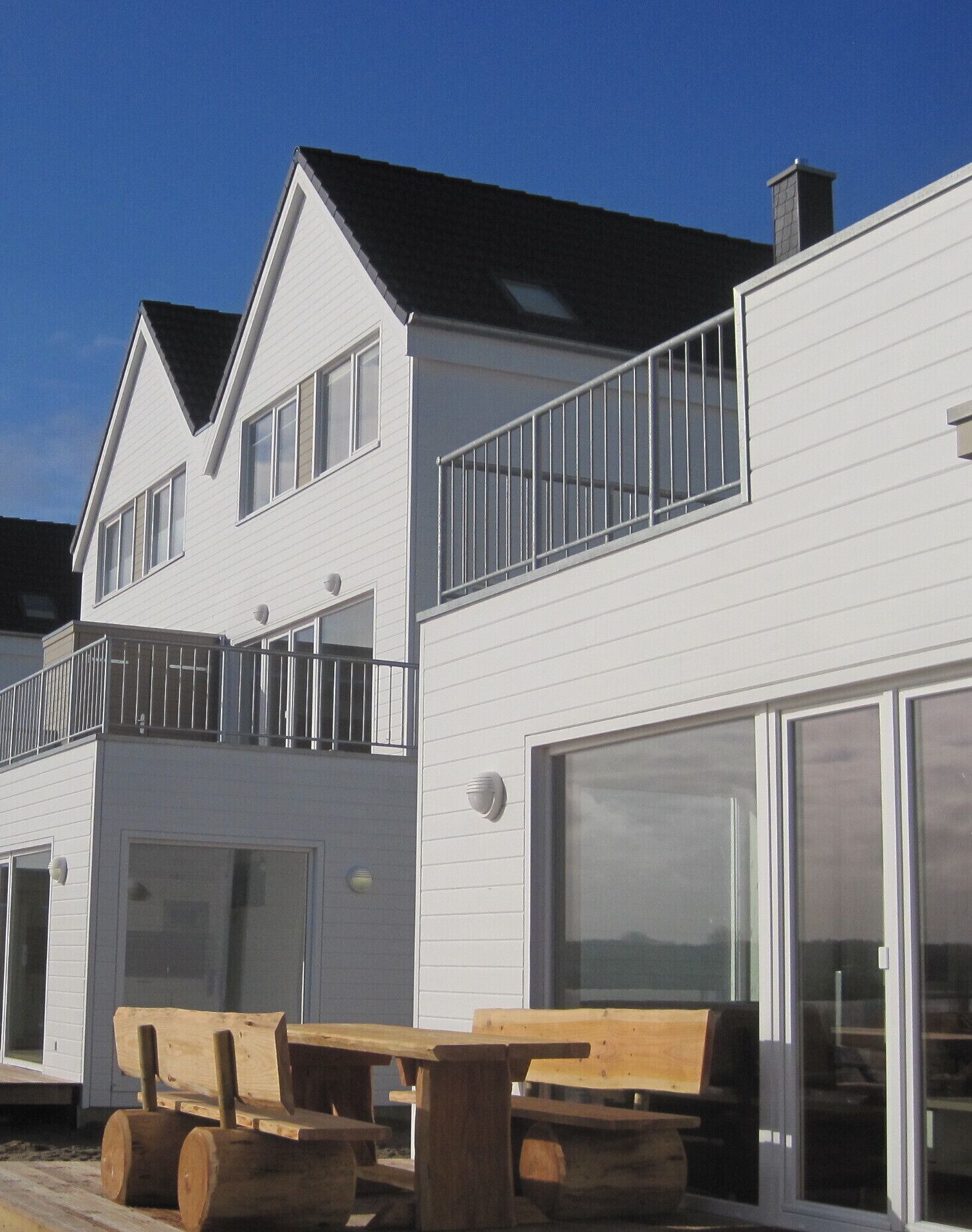 Een wit gebouw met twee verdiepingen, een balkon, een garage en een houten terras met bankjes.