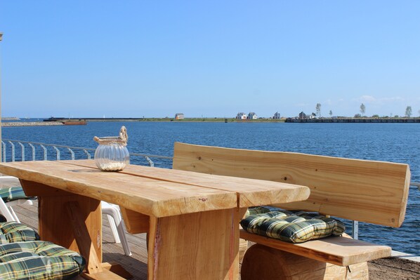 Une table en bois, un vase en verre et un banc agrémenté de coussins à carreaux, le tout installé au bord de l’eau.