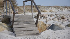 Una escalera de madera que sube hacia una plataforma con barandillas de metal, rodeada de rocas y arena.