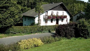 Une maison blanche avec un toit en pente, un balcon orné de jardinières et une haie au premier plan.