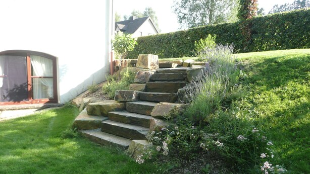 Un escalier en pierre menant à une maison, entouré d’une pelouse bien entretenue et de plantes en fleurs.