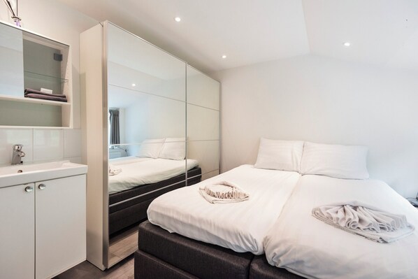A modern bedroom with a large mirror, a bed with white linens, and a built-in wardrobe.