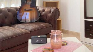 A living room with a brown leather sofa, a guitar, a Marshall amplifier, a vase with plants, and a pink mat with a pitcher of pink liquid.