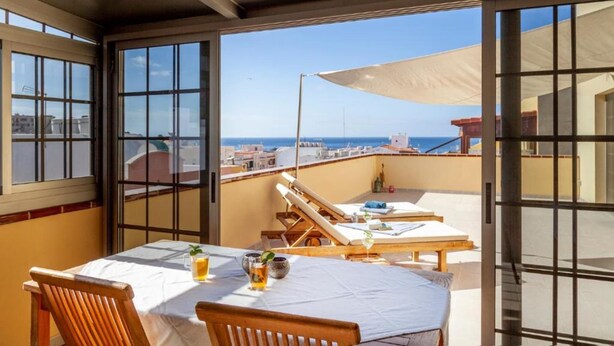 A balcony with a table set for two, a sun lounger, and a view of the sea.