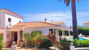 Una villa blanca con techo de tejas, un porche cubierto, piscina y una palmera.