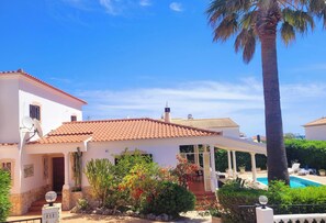 A white villa with a tiled roof, a covered patio, a swimming pool, and a palm tree.