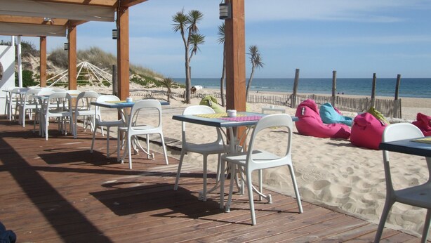 Un patio exterior con mesas y sillas, una terraza de madera y vistas a la playa y al océano.