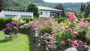 A stone wall with pink flowers in a garden area, a white building in the background, and a grassy lawn.