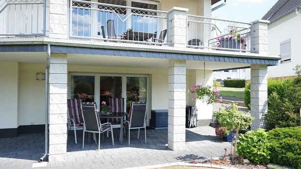 A two-story house with a covered patio area featuring a table and chairs, a balcony with a railing, and potted plants.