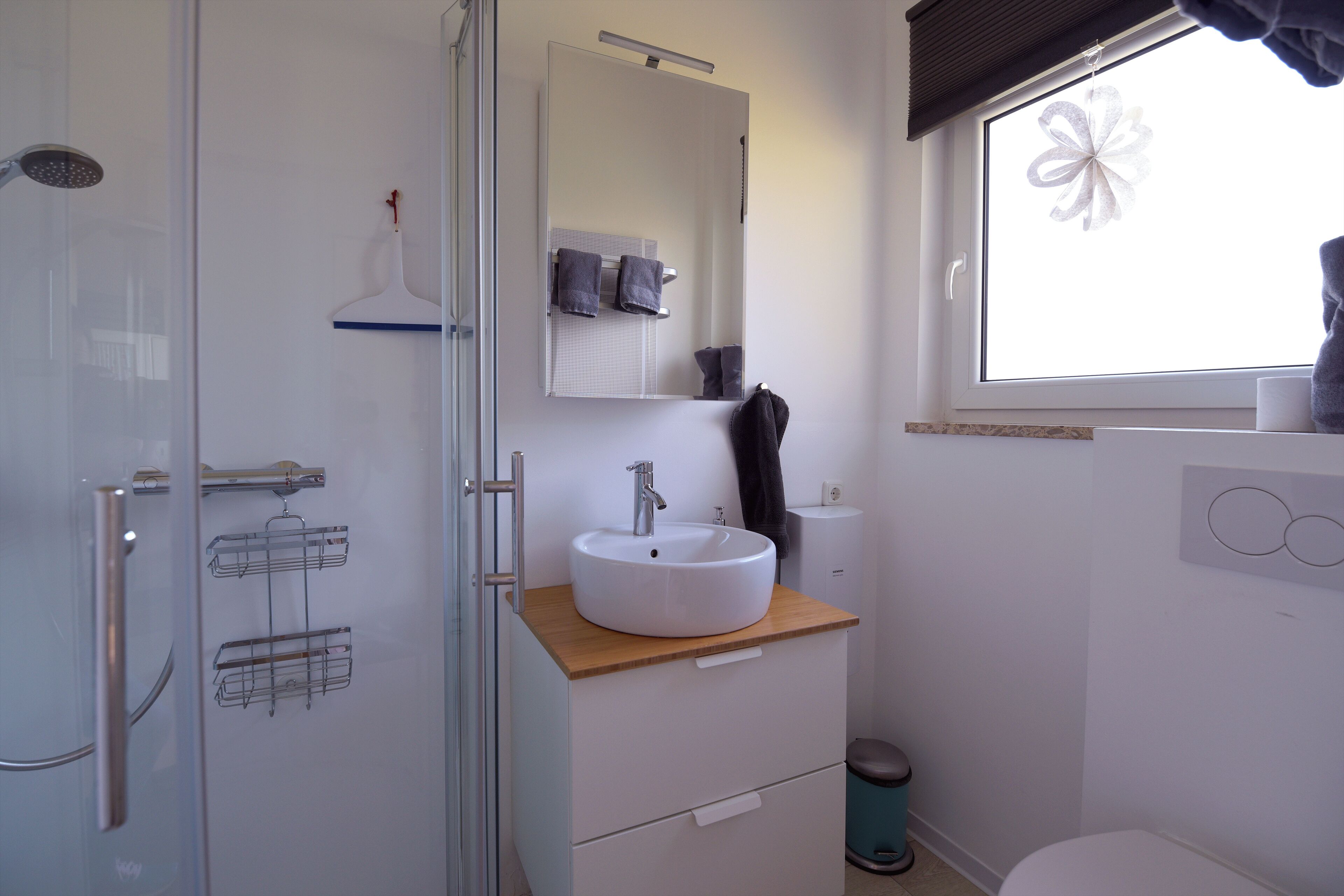 Ein modernes Badezimmer mit Duschkabine aus Glas, einem weißen Waschbecken mit Holzablage, einem Spiegel und einem Fenster mit floraler Dekoration.