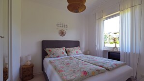 Een slaapkamer met een bed, een bedlampje, een klein nachtkastje, een raam met gordijnen en een wanddecoratie.