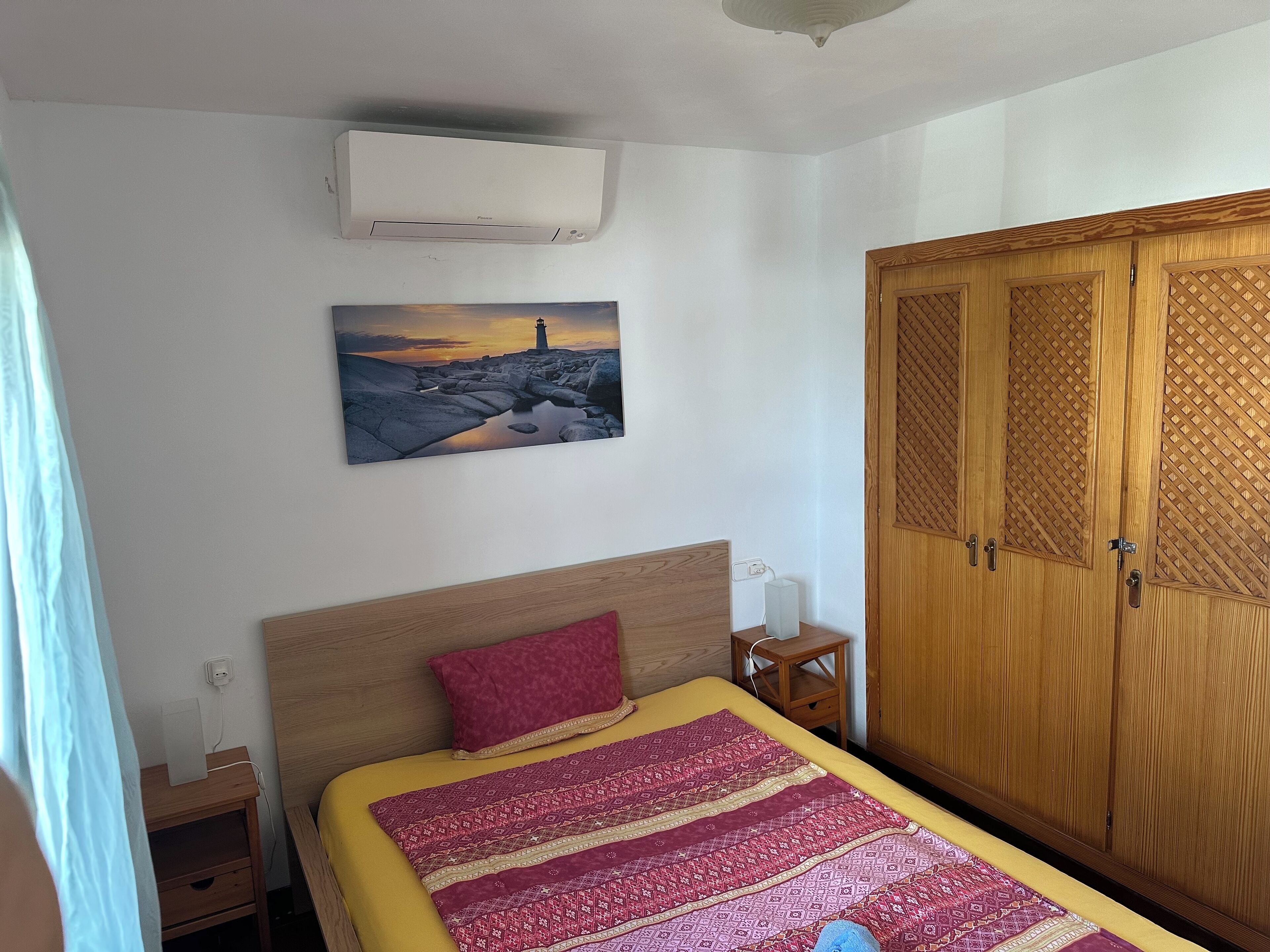 A bedroom with a bed, a nightstand, a wardrobe, an air conditioner, and a painting on the wall.