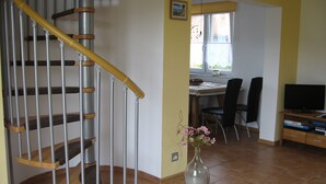 Un escalier avec des marches en bois et des rampes en métal, un coin repas avec une table et des chaises, un vase rempli de fleurs et un téléviseur posé sur un meuble.