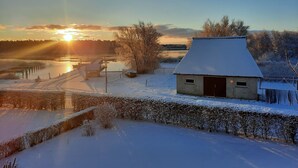 Un paysage enneigé avec une petite maison, un quai et une étendue d’eau, le tout baigné par les couleurs du coucher de soleil.