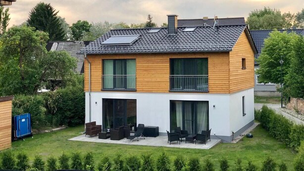 Ein modernes Haus mit zwei Obergeschossen, einer flachen Dachkonstruktion, großen Fenstern und einer Terrasse mit Gartenmöbeln.