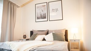 Ein ordentlich bezogenes Bett mit weißer Tagesdecke, einer Nachttischlampe und einem gerahmten Bild an der Wand.