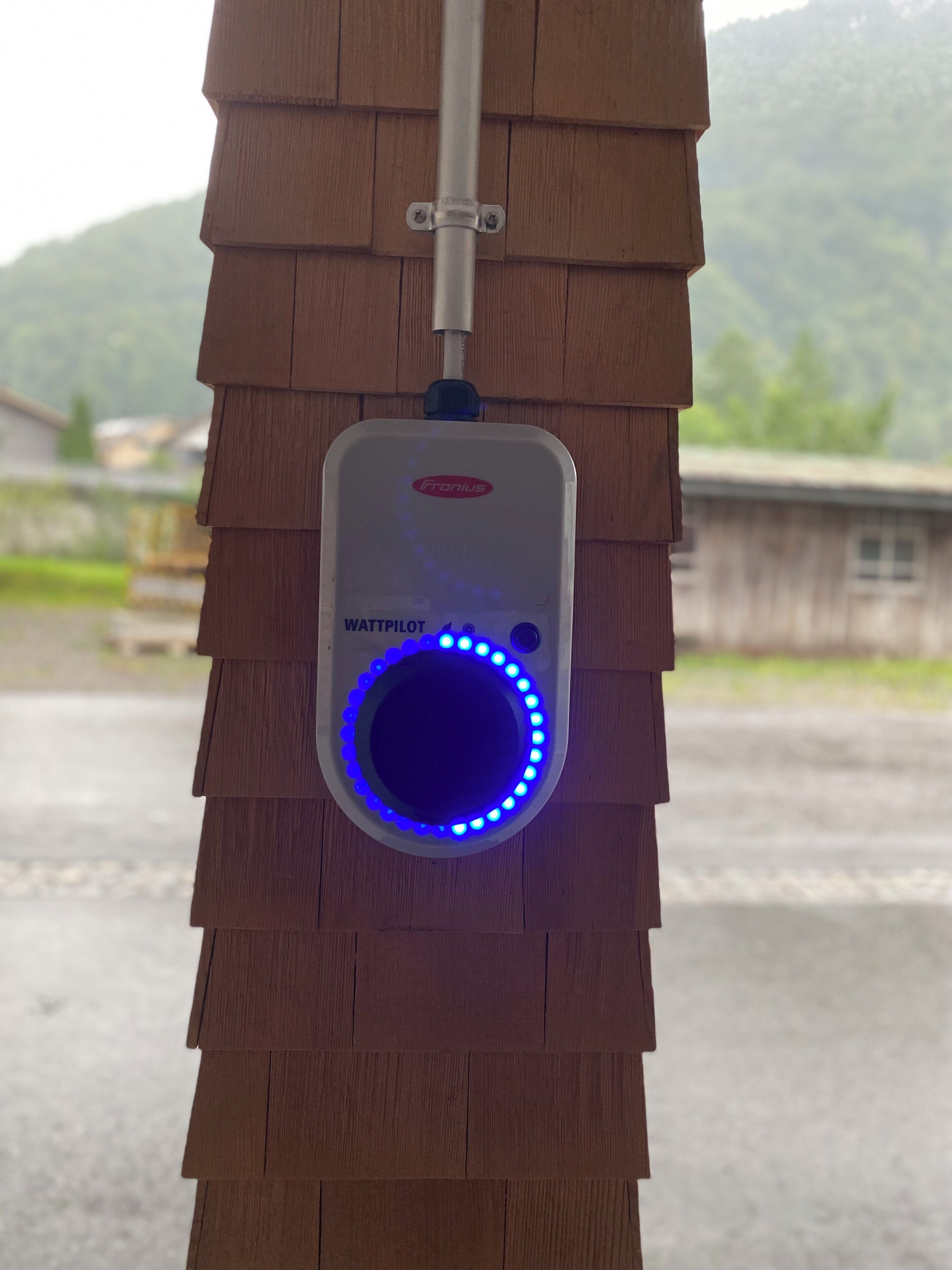 Ein Gerät mit der Aufschrift „WATTPILOT“, montiert an einer Holzkonstruktion, mit Blick auf eine ländliche Landschaft im Hintergrund.