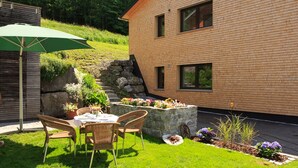 Une maison moderne en briques, dotée d’un patio comprenant une table et des chaises, le tout entouré d’un jardin bien entretenu, agrémenté de fleurs et de plantes.