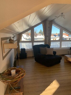 Un appartement-loft chaleureux, avec un canapé aux couleurs sombres, un plancher en bois et une grande fenêtre donnant sur le quartier.