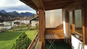 A wooden balcony with a view of a green lawn and houses, and mountains in the background.
