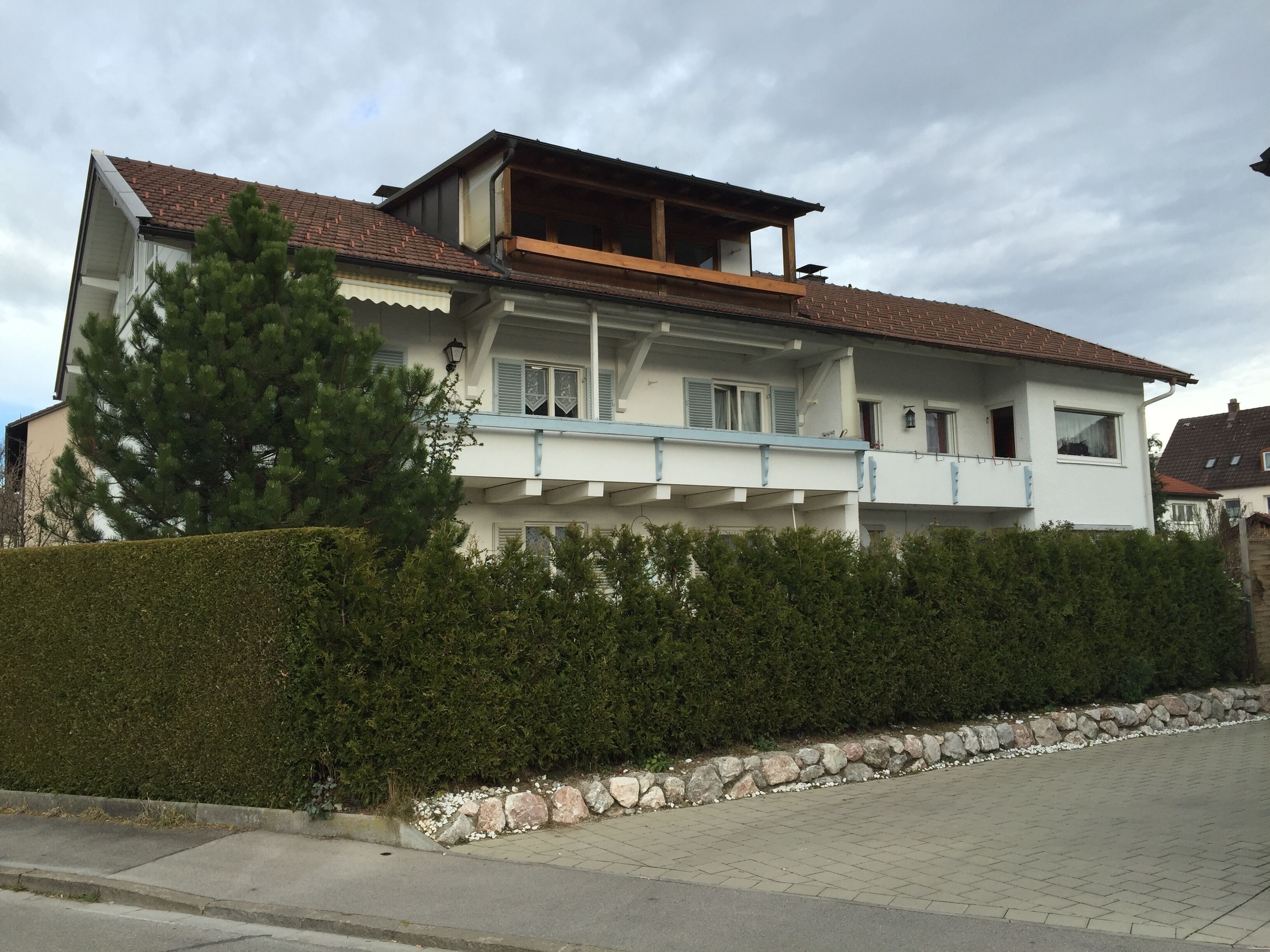 A two-story house with a balcony, a stone wall, and a hedge in front.