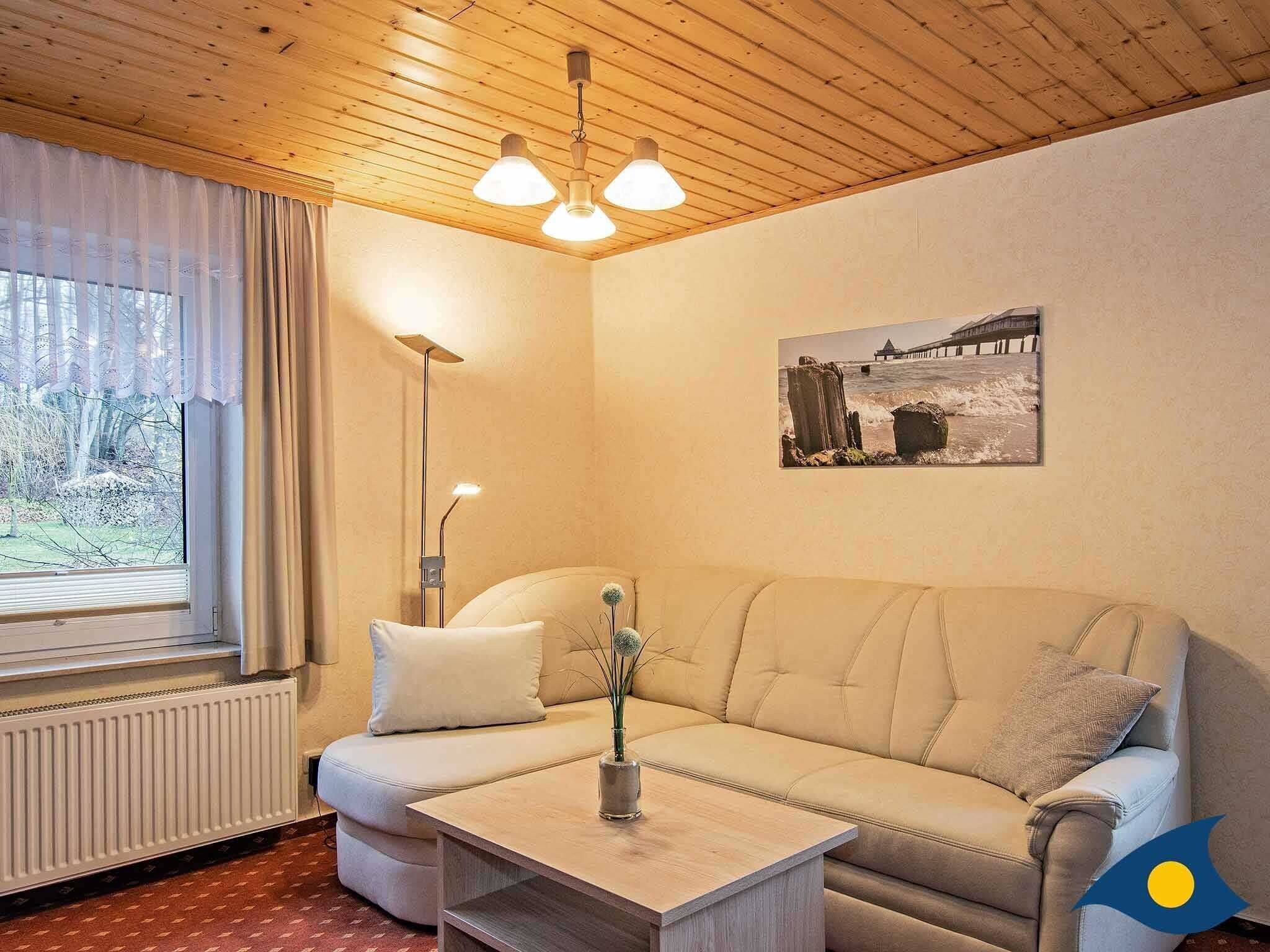 A cozy room with a wooden ceiling, a beige sofa, a small table with a vase, a floor lamp, and a window with curtains.