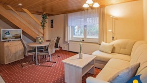 Ein gemütliches Zimmer mit Holzboden, einem Essbereich mit Tisch und Stühlen, einer Couch, einem Fernseher und einem Fenster mit Vorhängen.