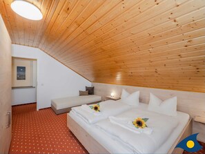 Un dormitorio con techo inclinado, una cama con ropa de cama blanca y girasoles, un pequeño sofá, una mesita de noche con lámpara, y un cuadro en la pared.