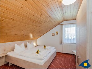 Une chambre avec un plafond en pente, un lit avec une literie blanche et des motifs de tournesols, une table de chevet avec une lampe, et une fenêtre avec des rideaux.