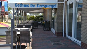 Ein Außenbereich mit Korbstühlen und Tischen, ein überdachter Bereich mit einem Schild, das "Eethuis De Zeespiegel" zeigt, und ein Pflasterweg, der zu einem Gebäude mit Glasüren führt.