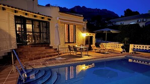 A swimming pool with steps leading up to a patio area, a covered seating area, and a view of mountains in the background.
