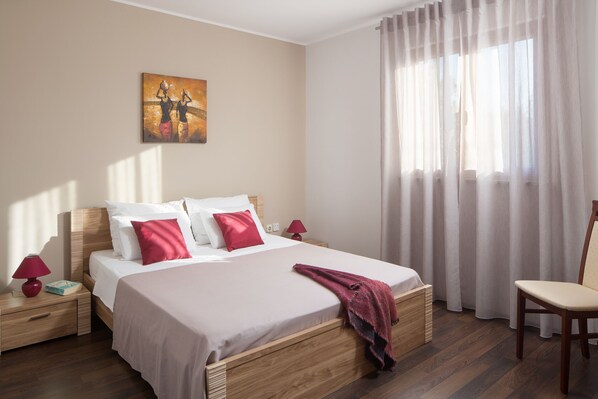 A neatly made bed with white and red bedding, a wooden headboard, and a red throw blanket. There is a nightstand with a red lamp and a book beside the bed. A painting hangs on the wall above the bed. - New luxurious and modern Villa, 5 minutes from the nearest beach (Krsan)