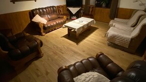 Una sala de estar con un sofá de cuero marrón, una mesa de centro de madera y un sillón color beige. Un televisor está montado en la pared, y hay una ventana con cortinas.