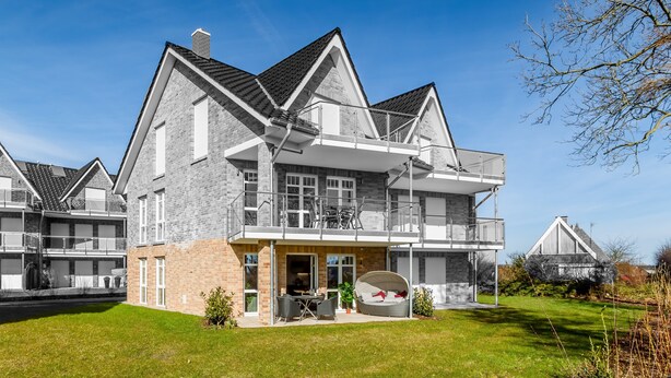 A modern two-story house with a balcony, a covered patio, and a well-maintained lawn. - Vacation home-Fehmarn (Fehmarn)