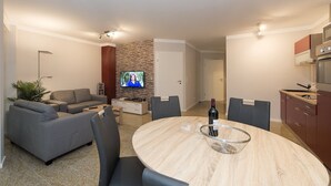 A modern living room with a gray sofa, a TV, a dining table with chairs, and a kitchen area with cabinets and a stove. - Vacation home-Fehmarn (Fehmarn)