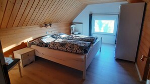 Une chambre avec un plafond en pente, un lit avec une literie à motifs, une table de chevet, une petite table, une fenêtre et un réfrigérateur.