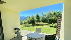 En terrasse med et rundt bord og to stole, et vindue, samt udsigt til et græsareal med træer og bjerge.