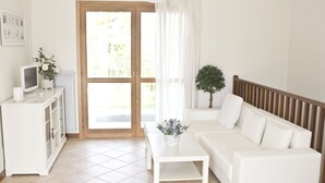 Ett modernt, väl upplyst rum med en vit soffa, ett litet vitt bord och en tv-apparat placerad på ett vitt skåp. Det finns en stor glasdörr som leder ut till ett utomhusområde.