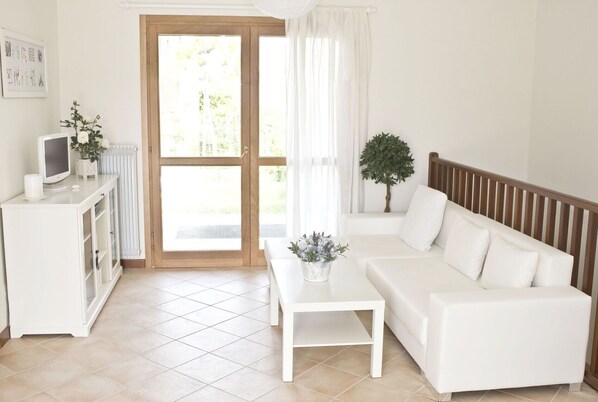 Une pièce moderne et bien éclairée, avec un canapé blanc, une petite table blanche et un téléviseur posé sur un meuble blanc. Une grande porte vitrée donne accès à un espace extérieur.
