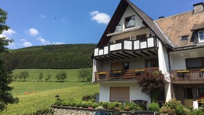 A two-story chalet with a balcony, surrounded by a lush green landscape and a forested hill. - Panorama-FeWo**** BEI WINTERBERG, 2 -10 pers., box-spring beds, WIFI / WLAN  (Schmallenberg)
