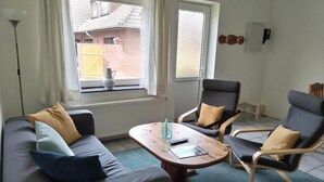 Ein Wohnzimmer mit einer Couch, zwei Sesseln, einem Couchtisch und Blick auf ein Backsteingebäude durch ein Fenster.