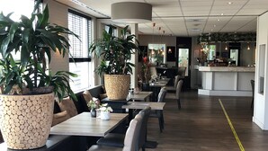 Un interno ristorante moderno con tavoli e sedie in legno, grandi piante in vaso e un ambiente pulito e ben illuminato.