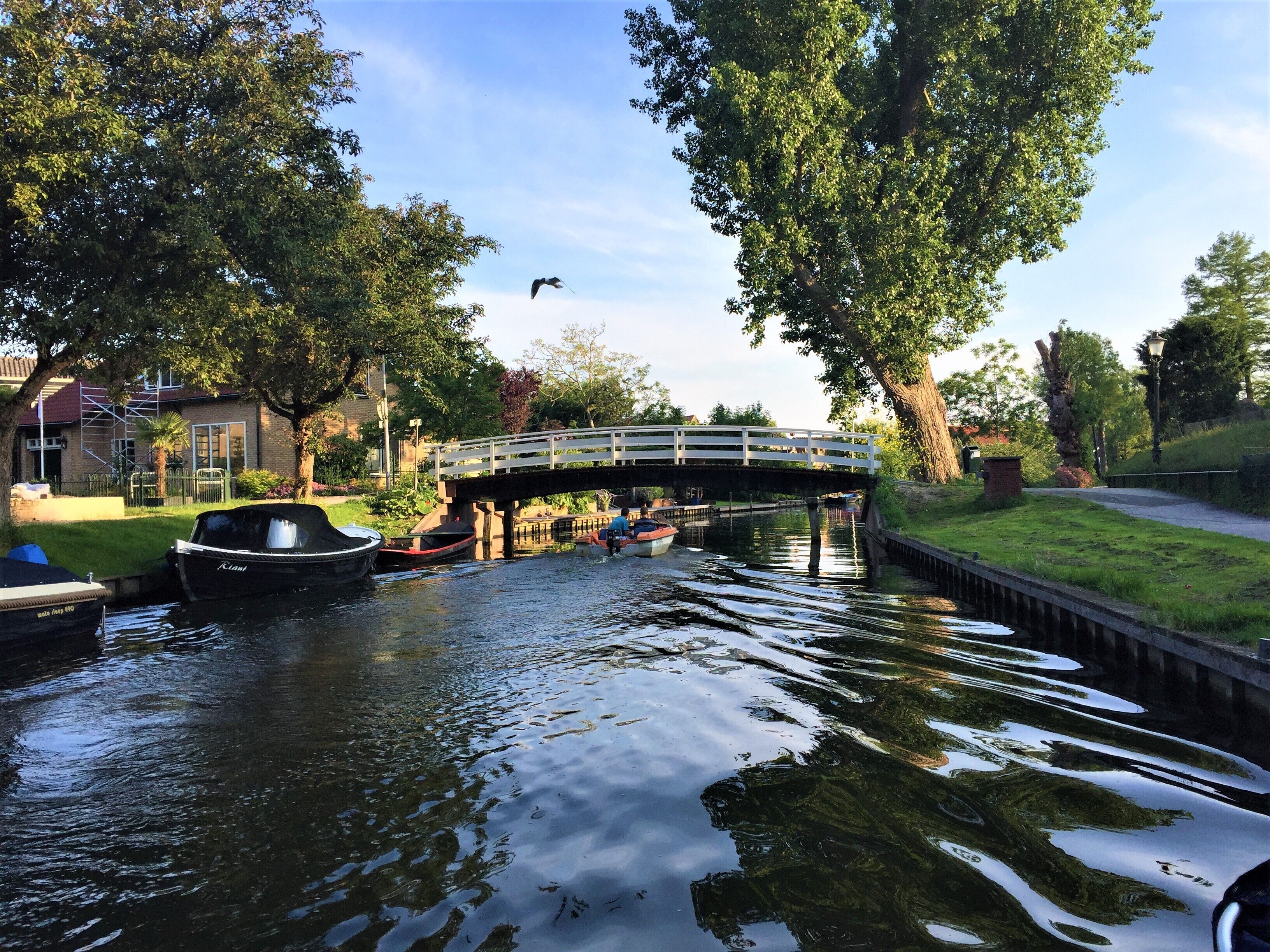 A canal with boats docked, a pedestrian bridge, and trees lining the water.