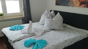 A neatly made bed with star-patterned bedding and towels, a window with curtains, a radiator, and a painting on the wall.