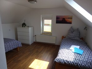 A small, well-lit room with a sloped ceiling, a radiator, a window with blinds, a bed with patterned bedding, a dresser, and a wall-mounted picture. - Vacation home Oll Tuun Rhauderfehn Leer Ostfriesland 6 pers. Fireplace infrared cabin (Rhauderfehn)
