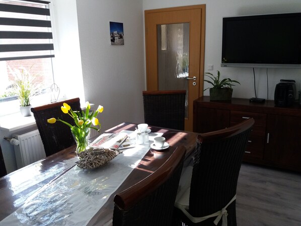 A dining area with a table set for two, a vase with yellow flowers, a newspaper, and a coffee maker.