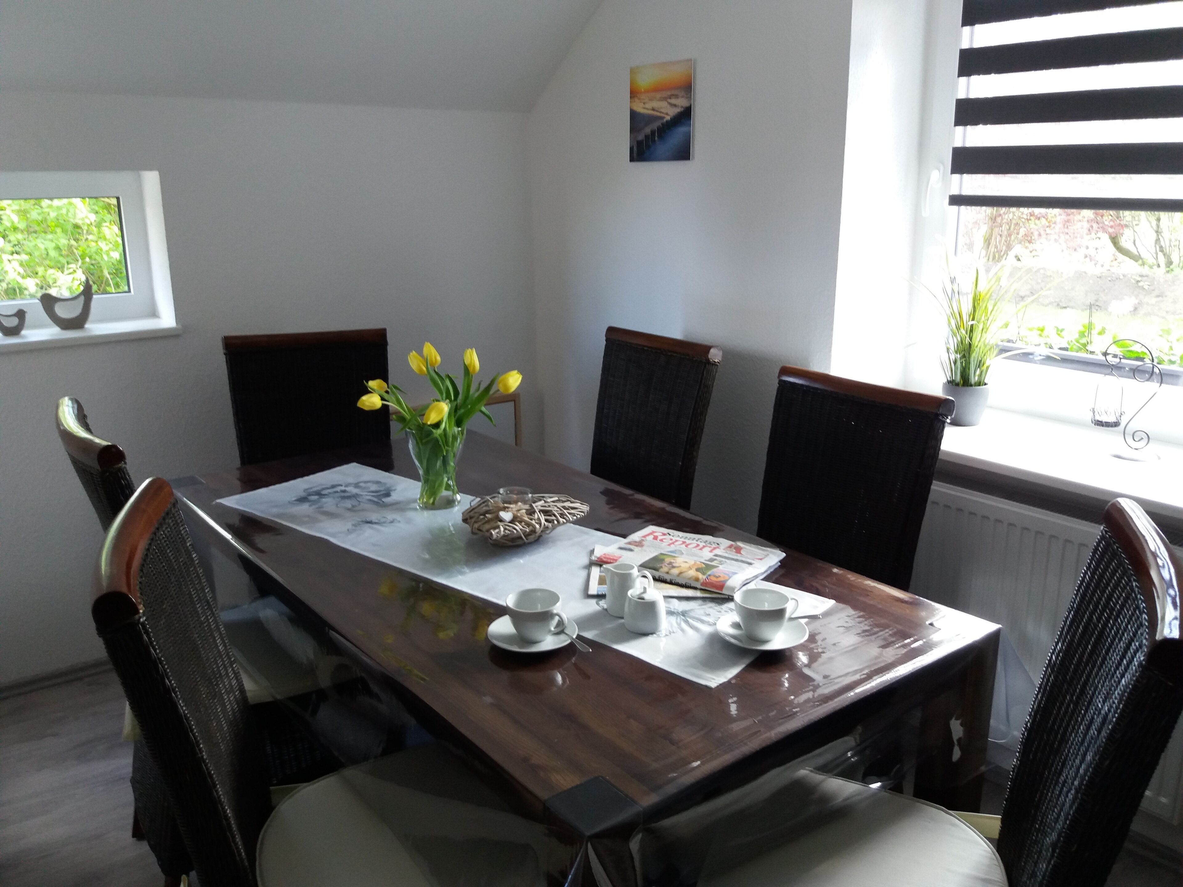 A dining area with a wooden table, chairs, a vase with yellow flowers, a magazine, and a window with a view of greenery.