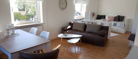 A modern living room with a dining area, wooden flooring, a brown sofa, a round clock, and large windows with blinds.