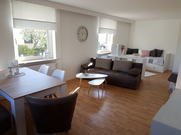 A modern living room with a dining area, wooden flooring, a brown sofa, a round clock, and large windows with blinds. - Modern, bright apartment in Waldeck - Höringhausen (Höringhausen)