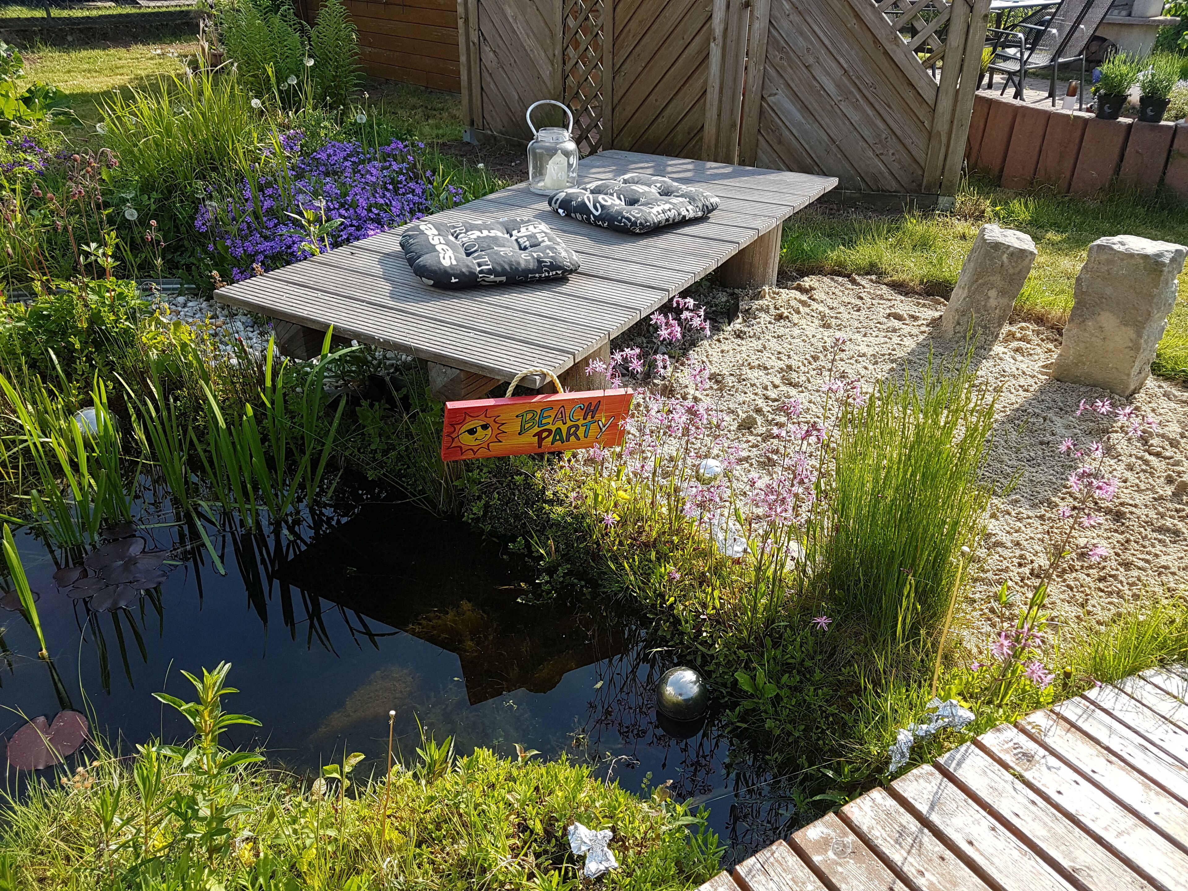 Een houten terras met een picknicktafel, omringd door een vijver, bloemen en een bord met de tekst 'Beach Party'.